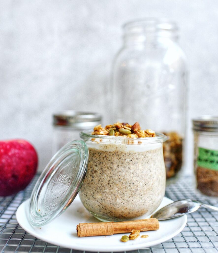 a glass jar filled with chia pudding. Granola is placed on top a cinnamon stick is placed on the plate below the jar. In the background is a blurred larger jar, apple and two smaller jars.