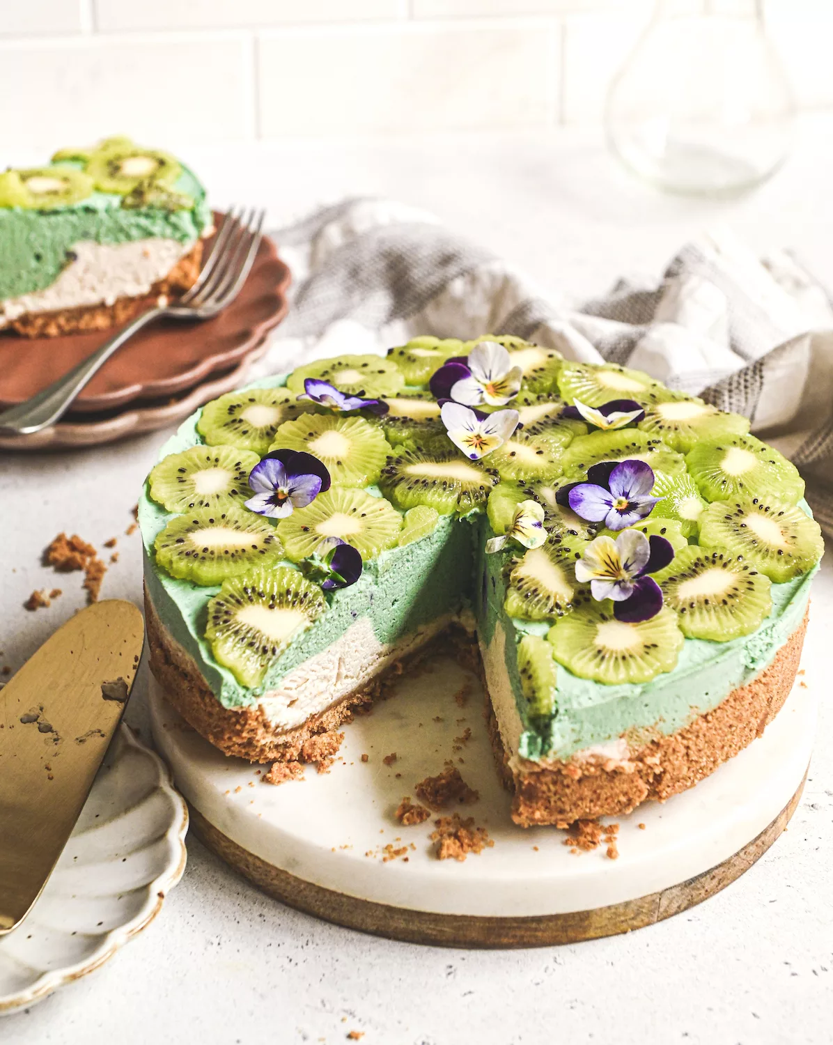 a beautifully decorated raw cheesecake with green and white layers, kiwi slices and edible flowers on top