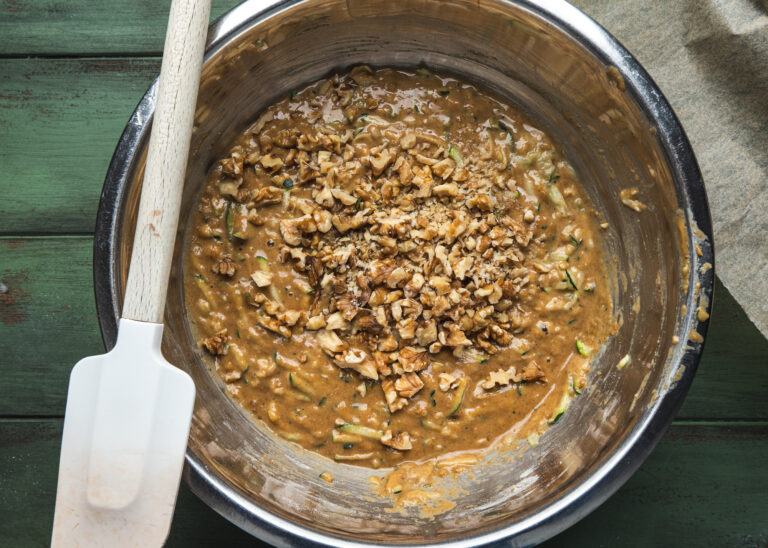 overhead view of silver bowl with wet mix of raw pecans, nuts butter, zucchini grated and spatula by teri-ann carty