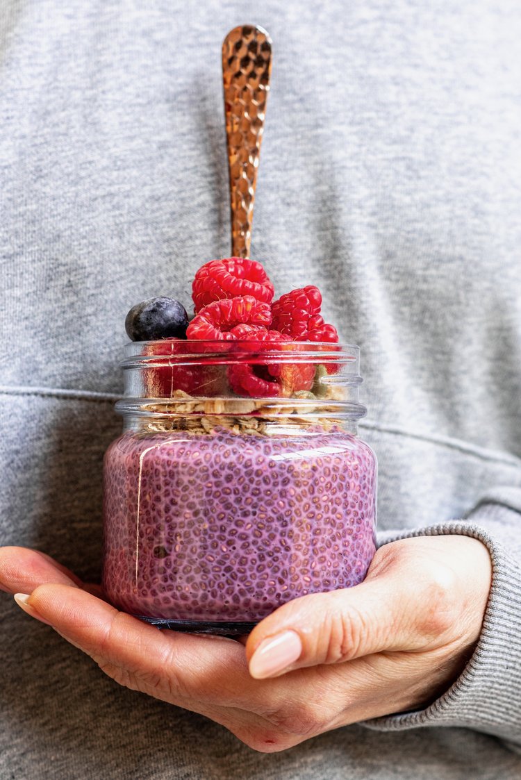 A small jar filled with purple chia pudding, raspberries and blueberries on top with a spoon. The jar is sitting in the hand of a female.