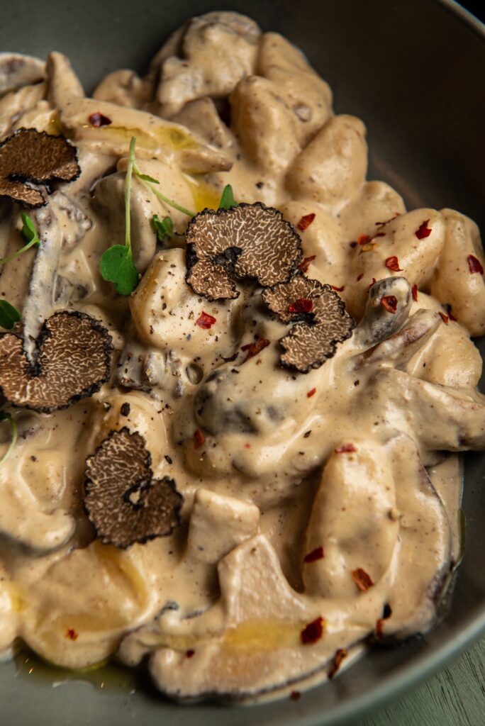 A close up image of Vegan BLACK TRUFFLE GNOCCHI pasta in a bowl. Thinly sliced Black truffle mushrooms are delicately placed on top.