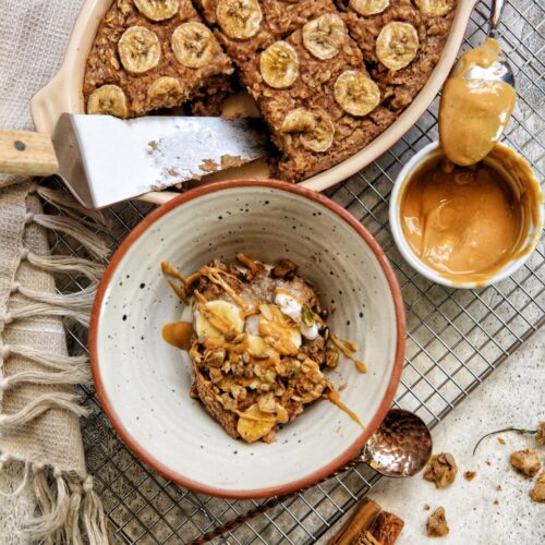 An arial view of a baking dish with one slice of banana baked oats cut out. a spoon covered in peanut butter placed slightly on the side of a bowl of peanut butter. there is also a bowl of the banana baked oats drizzled with peanut butter, banana slices and granola.