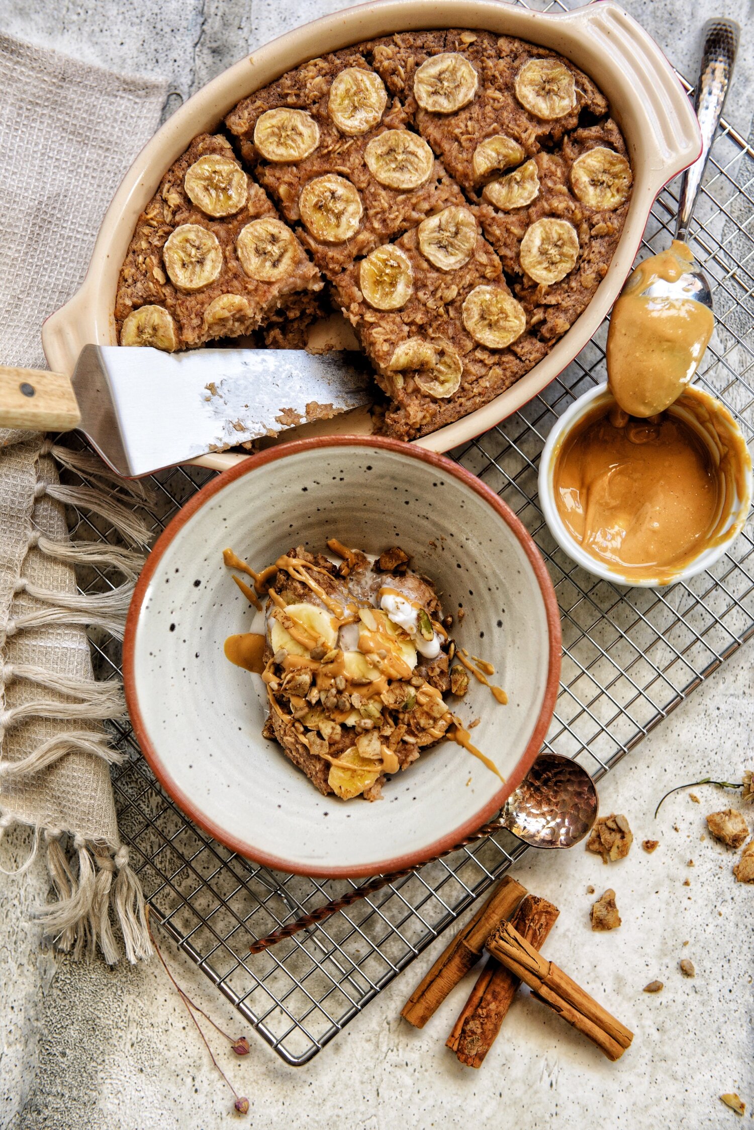 An arial view of a baking dish with one slice of banana baked oats cut out. a spoon covered in peanut butter placed slightly on the side of a bowl of peanut butter. there is also a bowl of the banana baked oats drizzled with peanut butter, banana slices and granola.