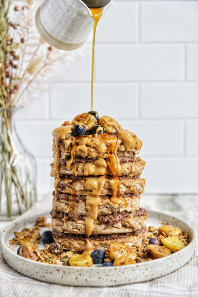 Delicious stack of gluten free, grain free, banana cassava pancakes with blueberries and fried banana on a plate and maple syrup being poured on top.