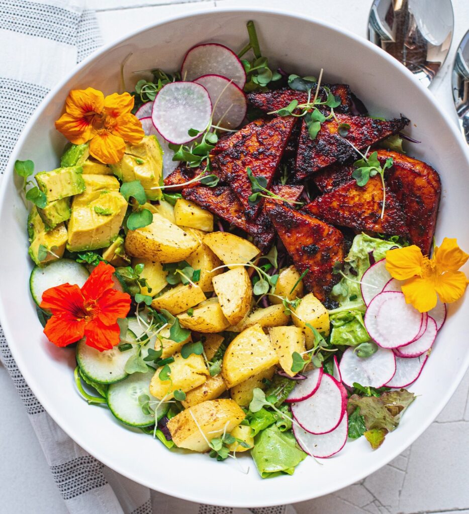 A beautiful salad bowl filled with colourful ingredients such as thinly sliced raddish, roasted potato, edible flowers, oven roasted bbq tofu, cubed avocado, sliced cucumber and micro greens