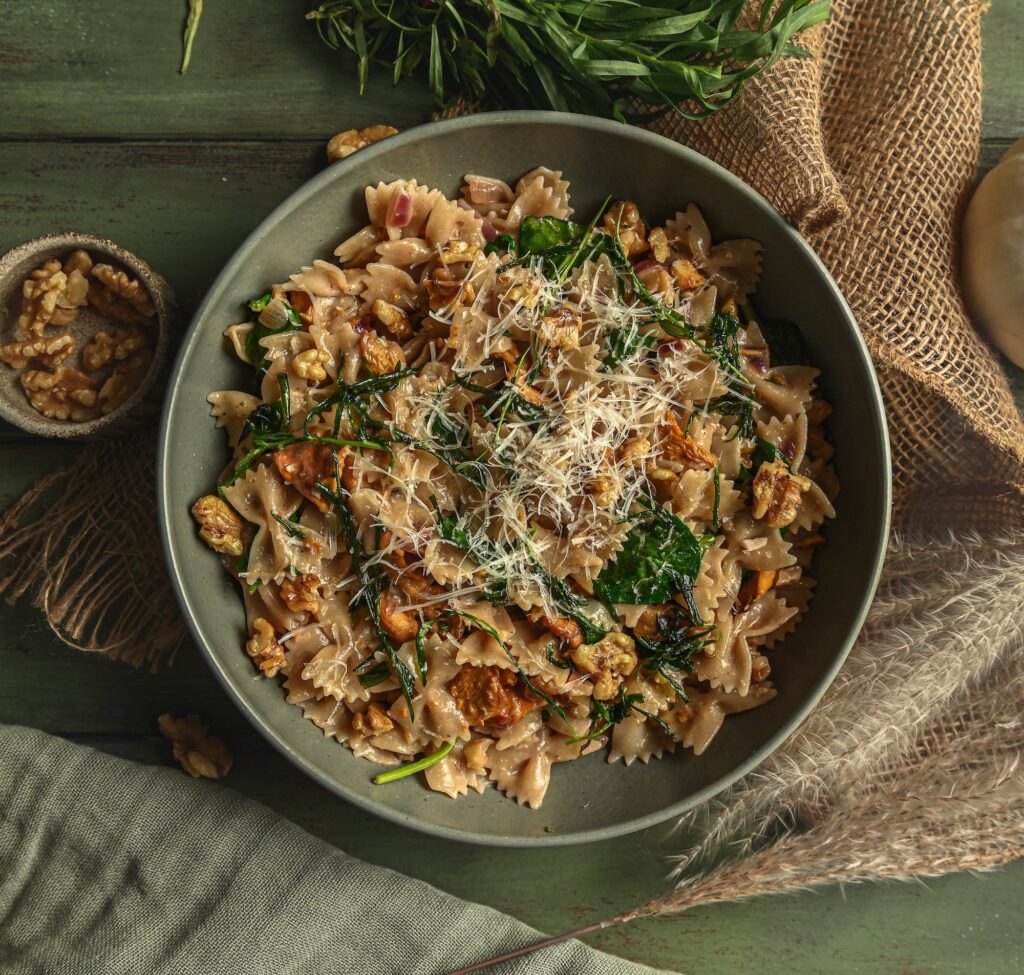 A bowl of Vegan Brown Butter Walnut and Tarragon Farfalle Pasta. A few raw walnuts scattered around the bowl and freshly grated vegan parmesan cheese on top.