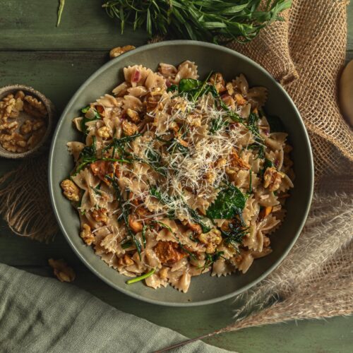 A bowl of Vegan Brown Butter Walnut and Tarragon Farfalle Pasta. A few raw walnuts scattered around the bowl and freshly grated vegan parmesan cheese on top.
