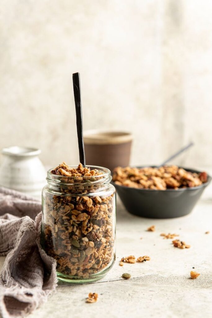 a jar with granola inside, in the background is a blurred vision of a black bowl with more granola and two pottery pots