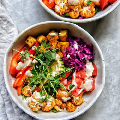 two bowls of beautifully colourful vegetables like microgreens, red pepper, cauliflower, tofu, cabbage and tahini dressing drizzled over all the ingredients.