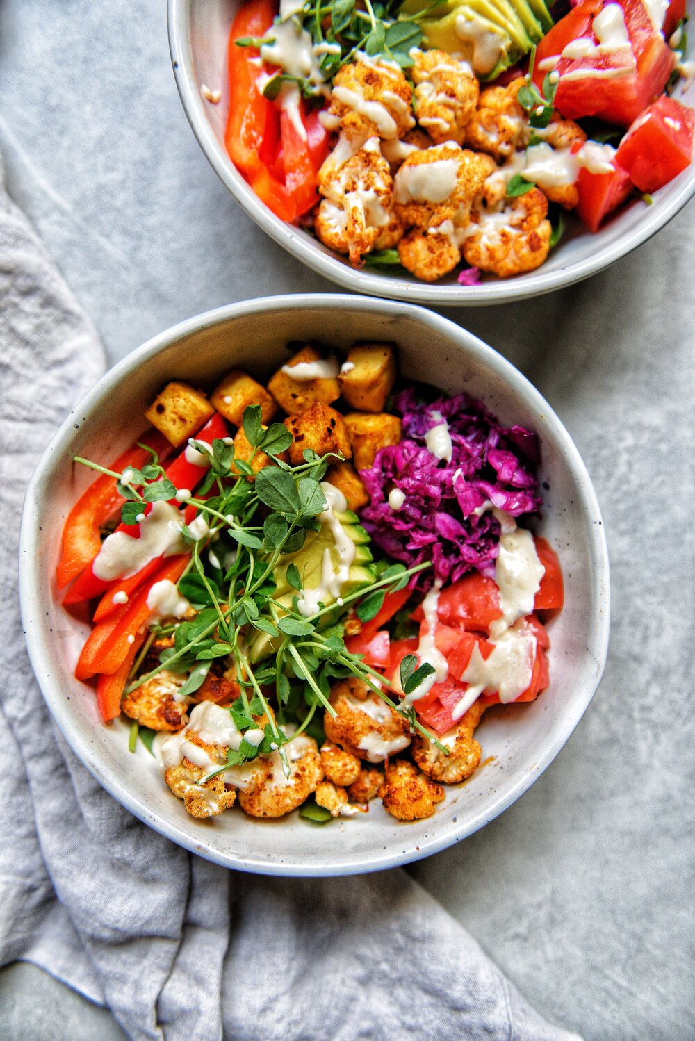 two bowls of beautifully colourful vegetables like microgreens, red pepper, cauliflower, tofu, cabbage and tahini dressing drizzled over all the ingredients.