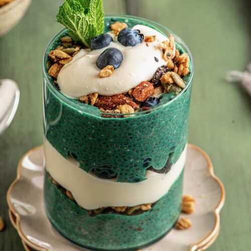vegan yogurt and green chia pudding layered in a clear glass with granola blueberries on top. sitting on a scallop trimmed plate with gold rim and a fresh sprig of mint on top.