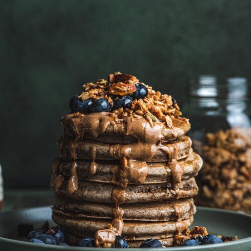 a seven pancake stack on a plate with peanut butter, blueberry and granola on top.
