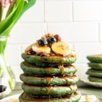 a stack of green pancakes on a plate with a drizzle of maple syrup from the top, bananas, granola and blueberries on the plate and the top of the pancake stack.
