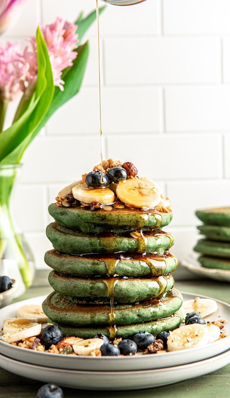 a stack of green pancakes on a plate with a drizzle of maple syrup from the top, bananas, granola and blueberries on the plate and the top of the pancake stack.
