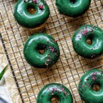 cooling rack with freshly baked, green glazed donuts and flakes of rose petals by teri-ann carty