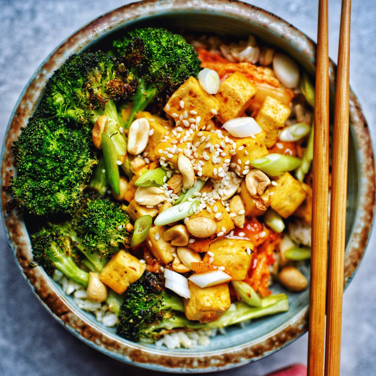 bowl of tofu, broccoli, peanuts with wooden chopsticks on top by teri ann-carty