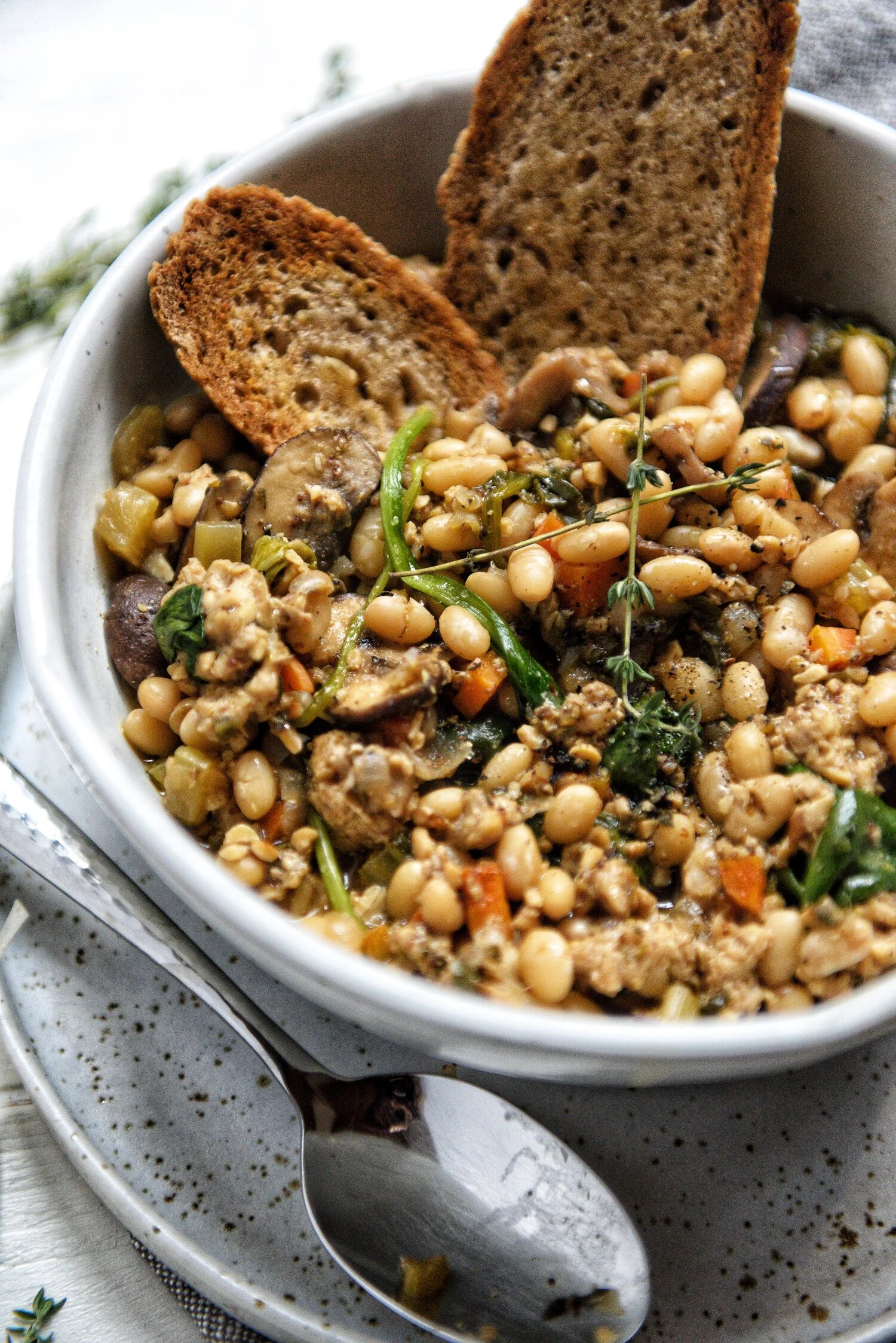 a beautiful ceramic dish filled with a white bean cassoulet. There is toasted bread on the side of the dish with a spoon and some greens on top
