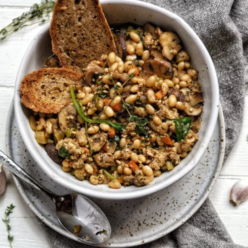 a beautiful ceramic dish filled with a white bean cassoulet. There is toasted bread on the side of the dish with a spoon and some greens on top