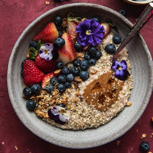 a beautiful ceramic bowl with steel cut oats, a sprinkle of seeds, blueberries and freshly sliced strawberries and edible purple flowers