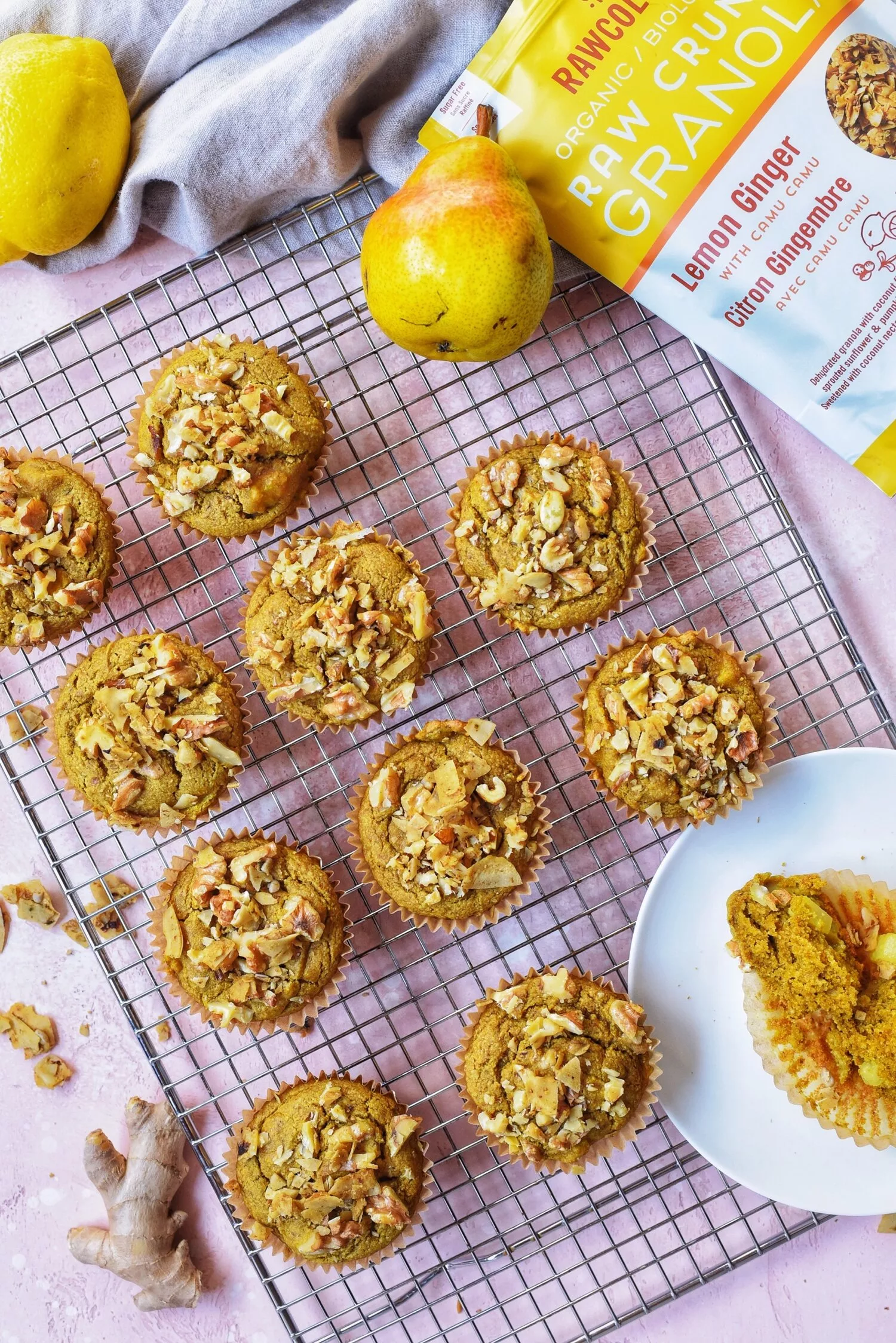 a cooling tray filled with beautifully decorated yellow turmeric lemon muffins, accompanied by a pear and bag of rawcology granola