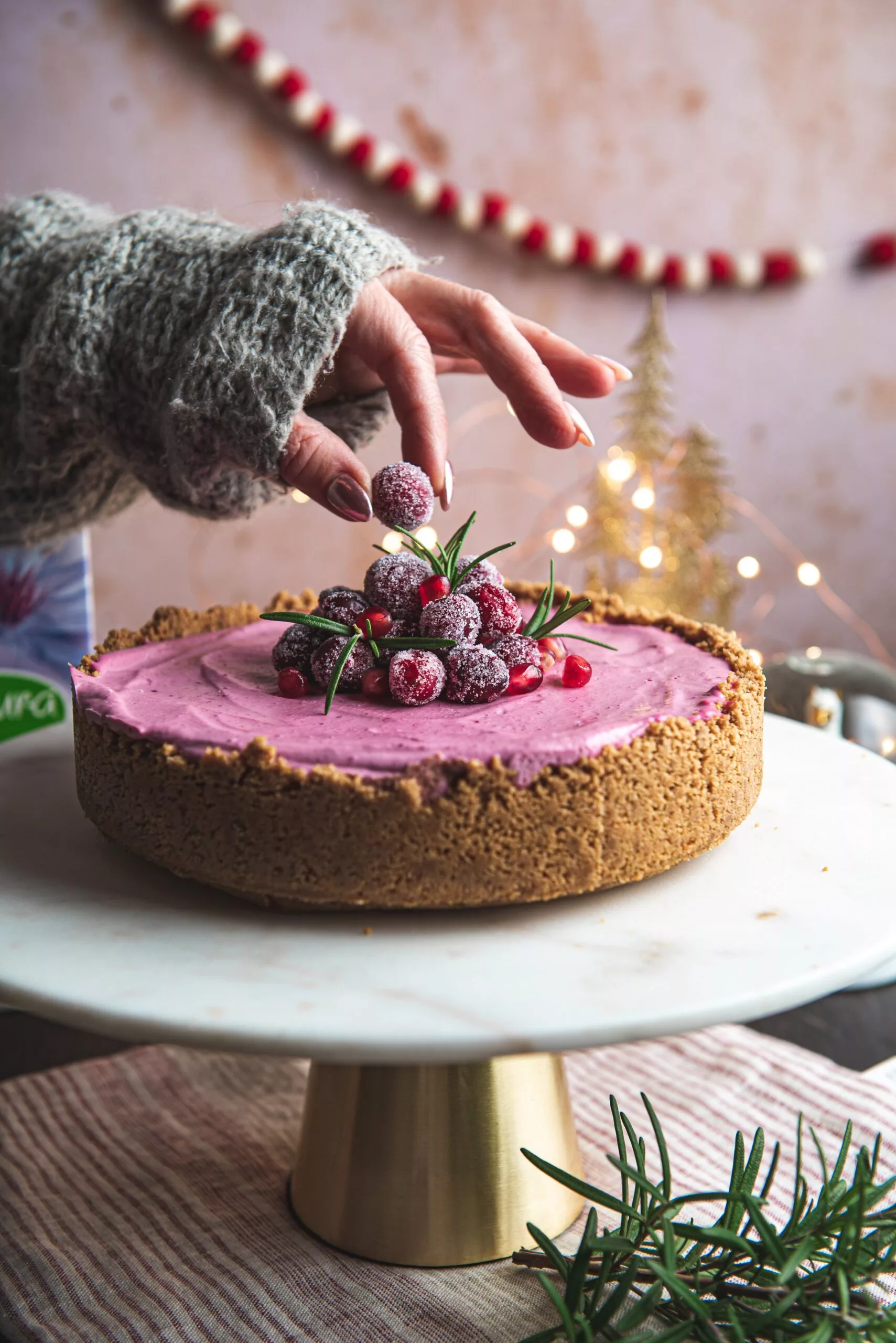 pink cheesecake with sugared cranberries and festive decorations. person placing sugared berry on top of cake