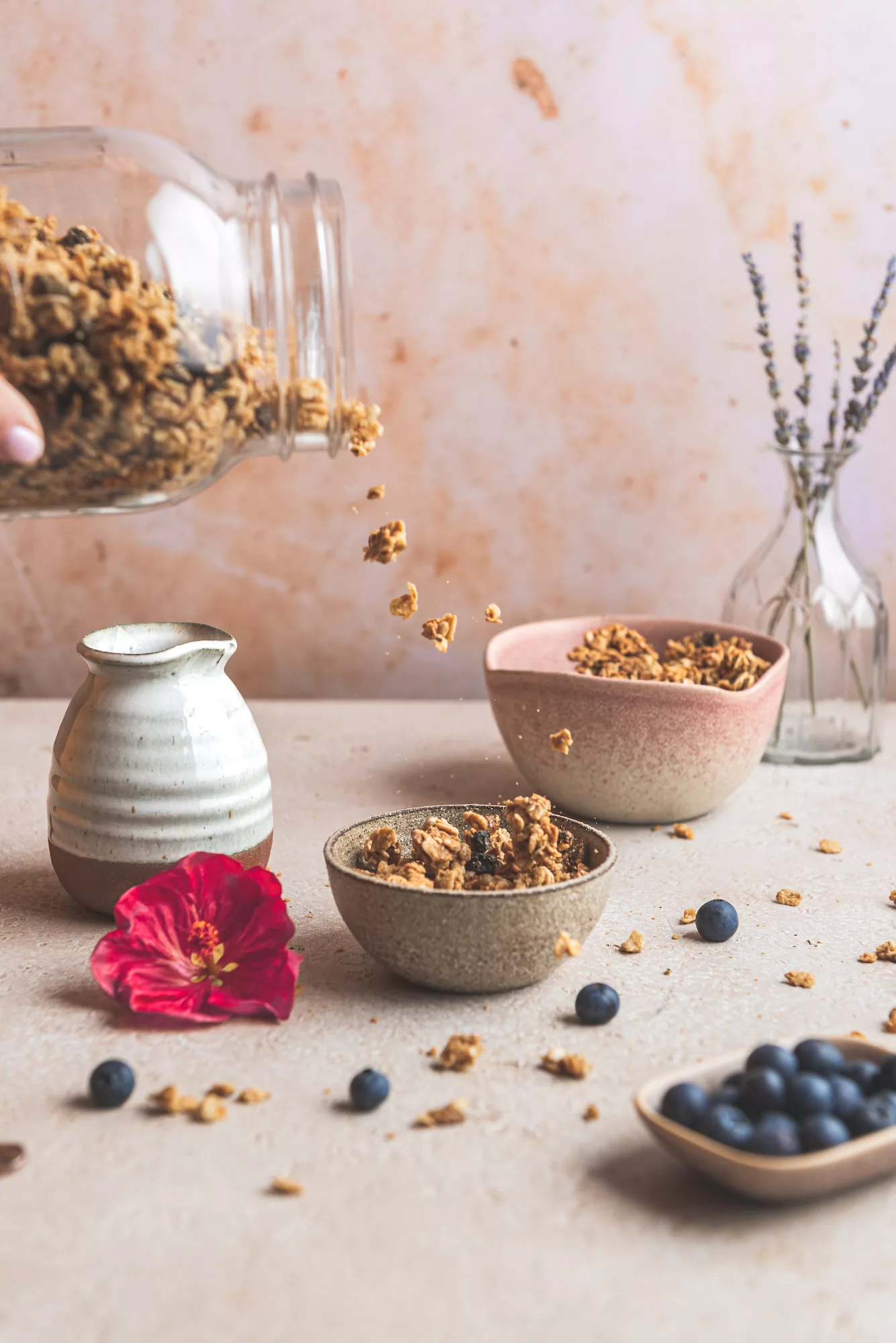 granola being poured into a bowl of granola, blueberries and hibiscus on the table around the bowl