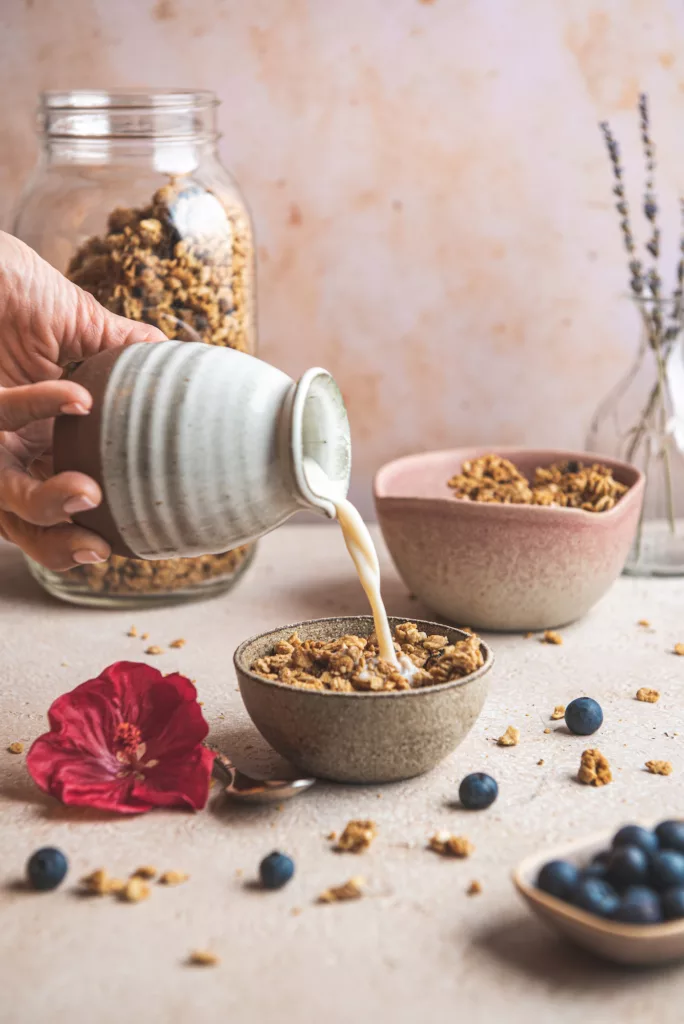 milk being poured into a ceramic bowl filled with granola, hibiscus flower on the side and blueberries as well.