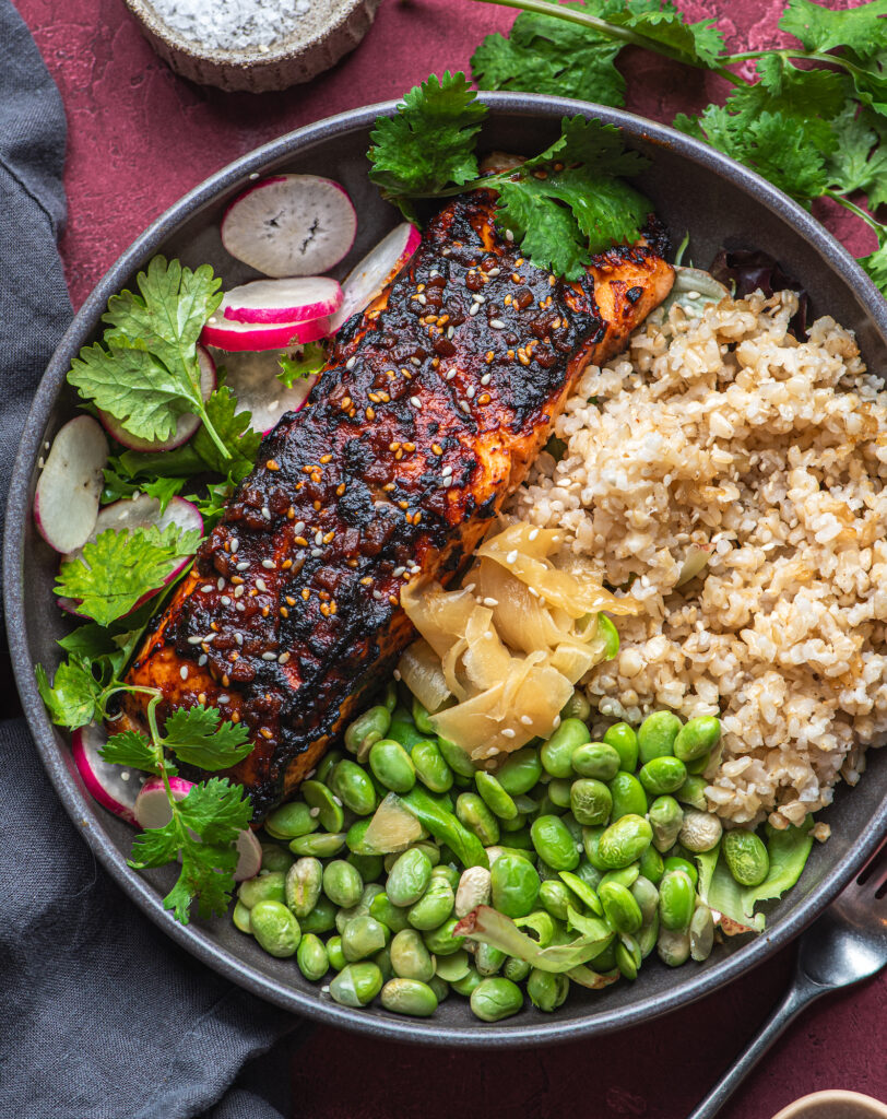 overhead view of healthy bowl with brown rice, edamames, cilantro, radish and crispy crusted salmon by teri-ann carty
