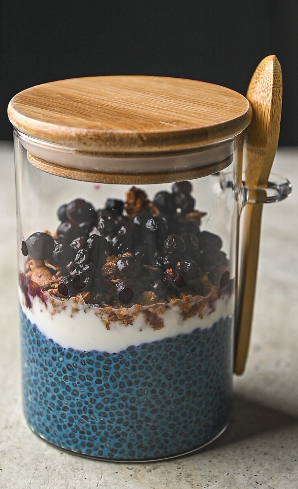 Glass jar with wooden lid and spoon, inside contains overnight blue chia pudding, granola and blueberries ready to go by teri-ann carty