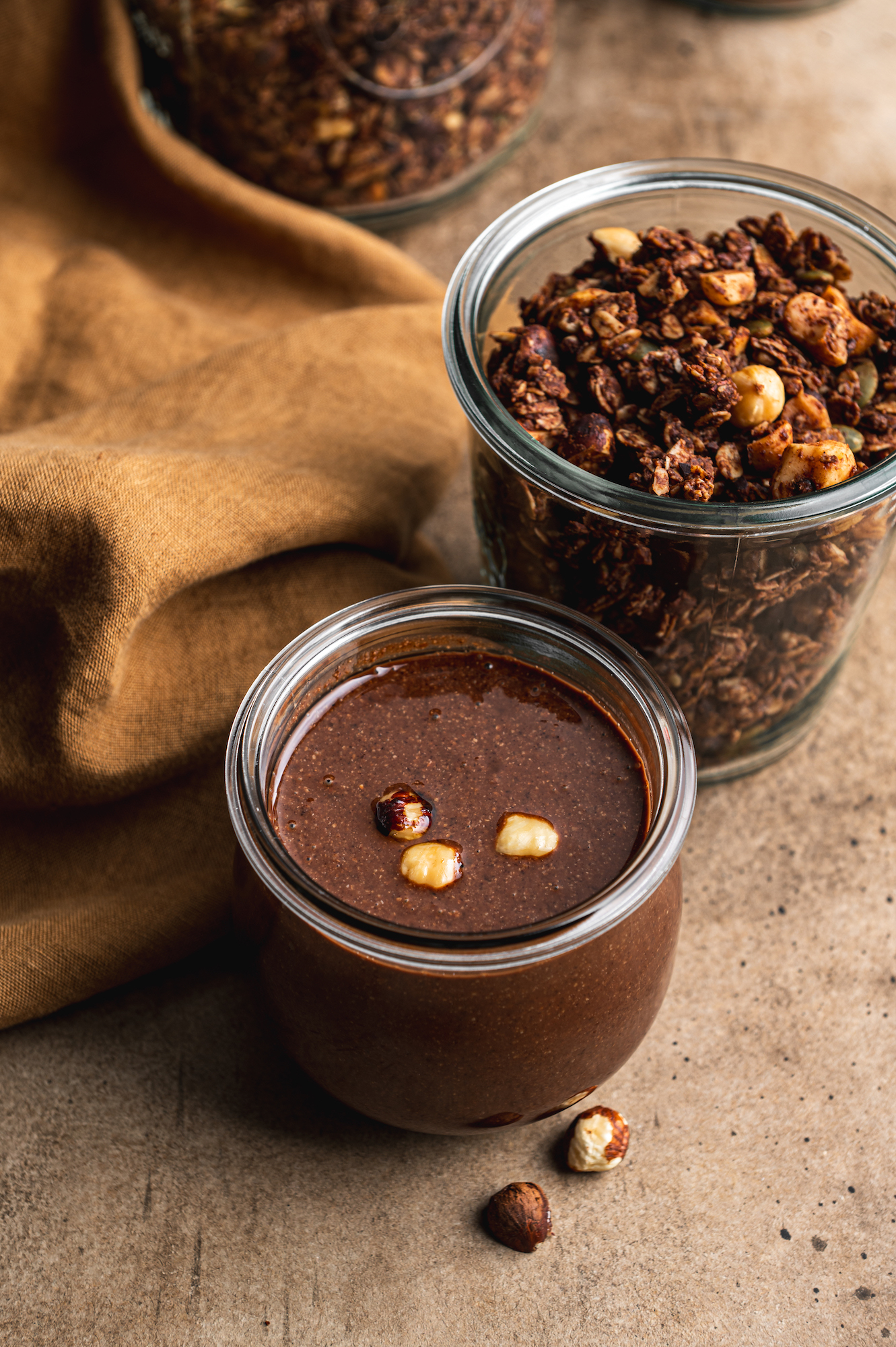 glass jar filled with freshly homemade chocolate nutella and a jar with chocolate granola behind by teri-ann carty