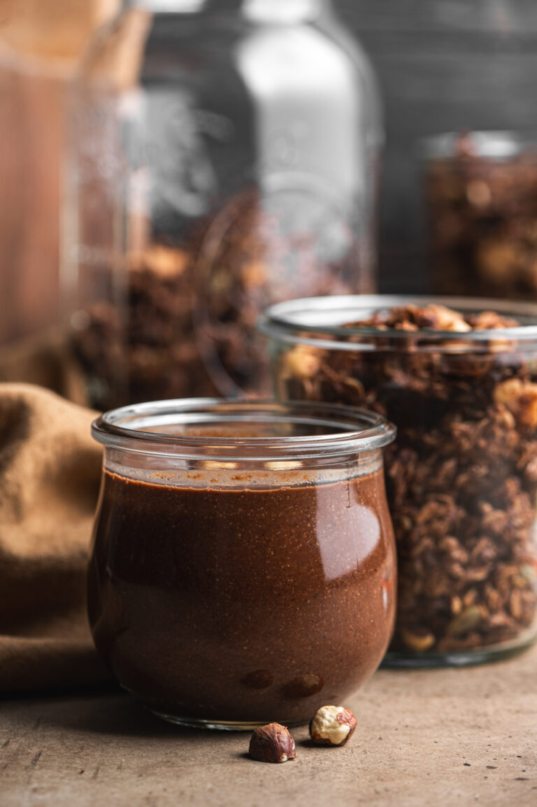 glass jar filled with freshly homemade chocolate nutella and a jar with chocolate granola behind by teri-ann carty