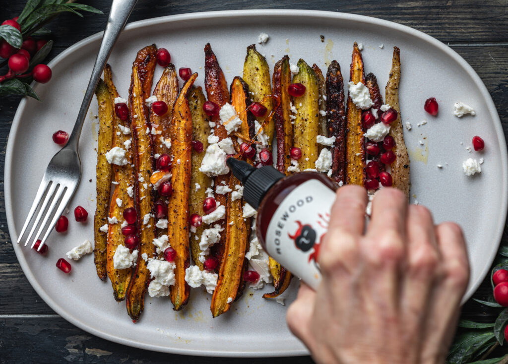 hand adding hot honey to a freshly roasted carrots decorated with cheese and baked seeds by teri-ann carty