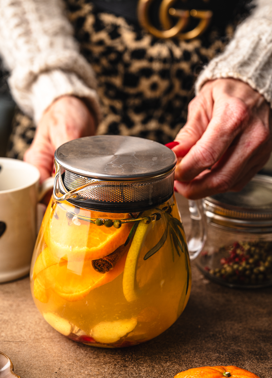 hands holding a glass teapot filled with citrus tea, rosemary, cinnamon and whole spices