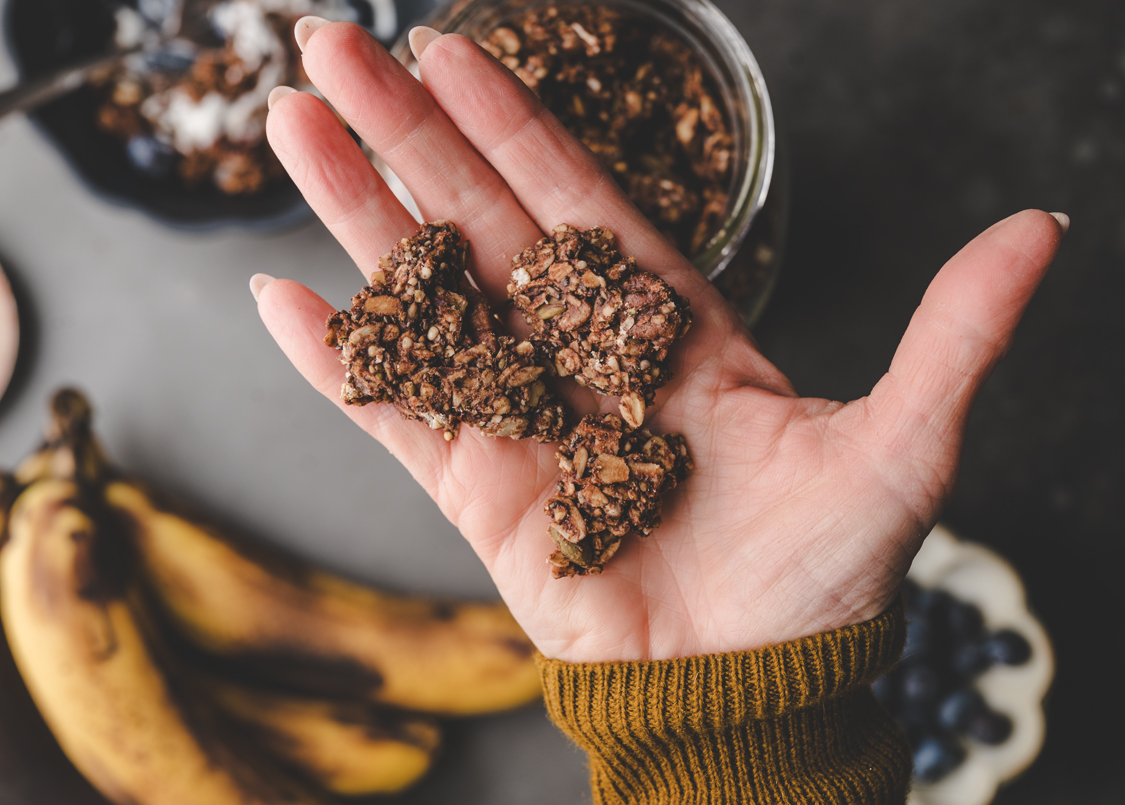 A hand holding pieces of granola and bananas in the background