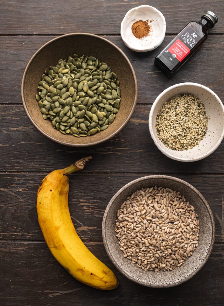 banana alongside bowl of pumpkin seeds, sunflower seeds, hemp seeds, cinnamon and vanilla extract all laid out on a wood table