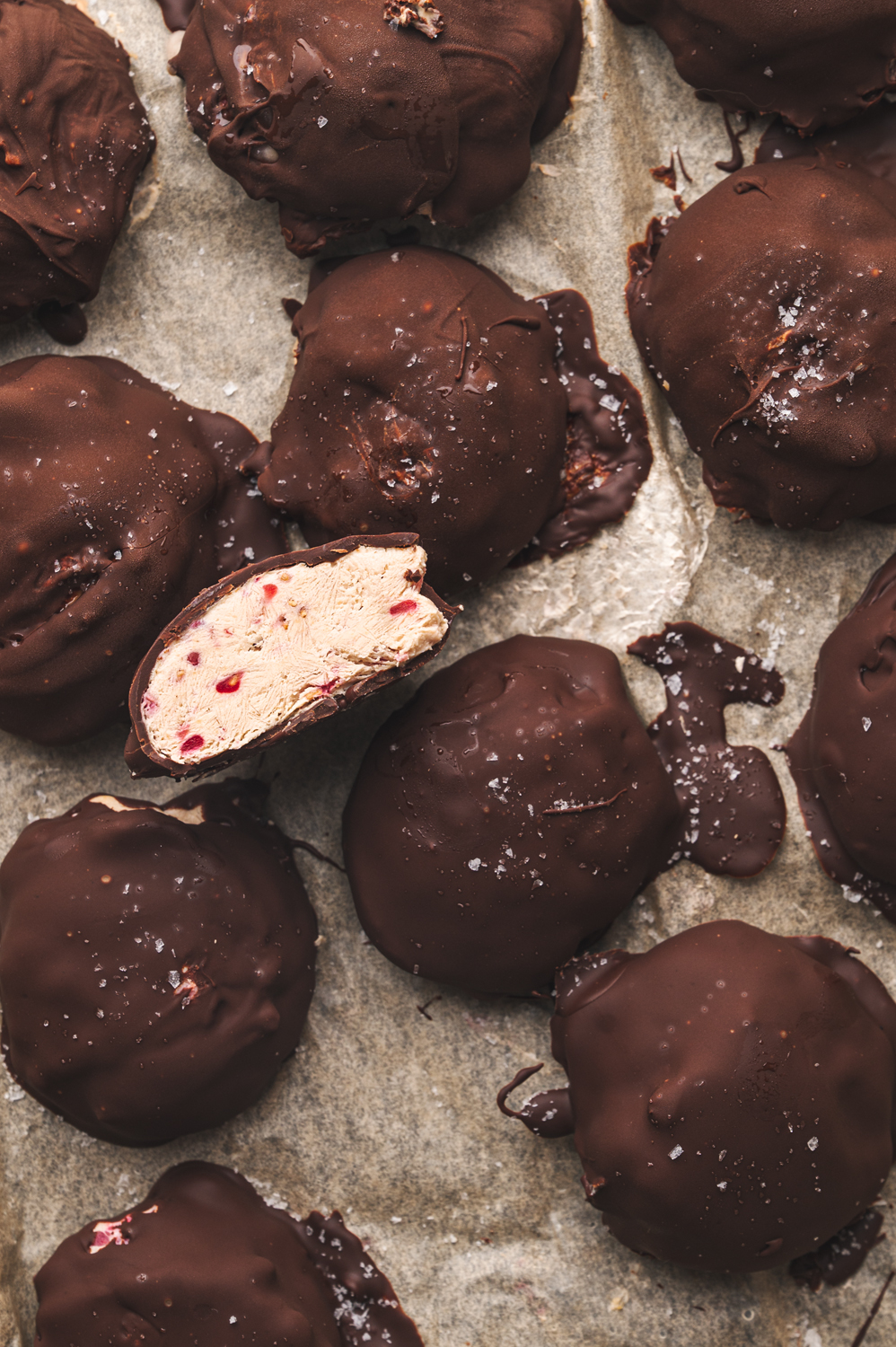 parchment paper with several chocolate covered yogurt bites sprawled out, one yogurt bite is cut open to show the delicious inside