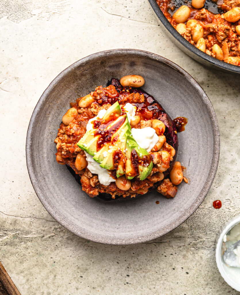 large ceramic bowl filled with high protein stuffed sweet potatoes loaded with ground beef, white beans, avocado, cottage cheese and a drizzle of chili cruch oil
