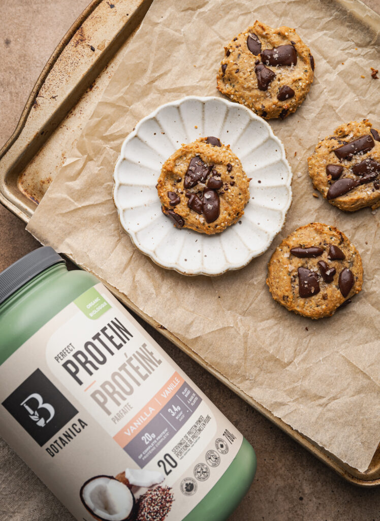 botanica vanilla protein powder container alongside a baking sheet with four chocolate chip protein cookies, one cookie is on a white scalloped plate