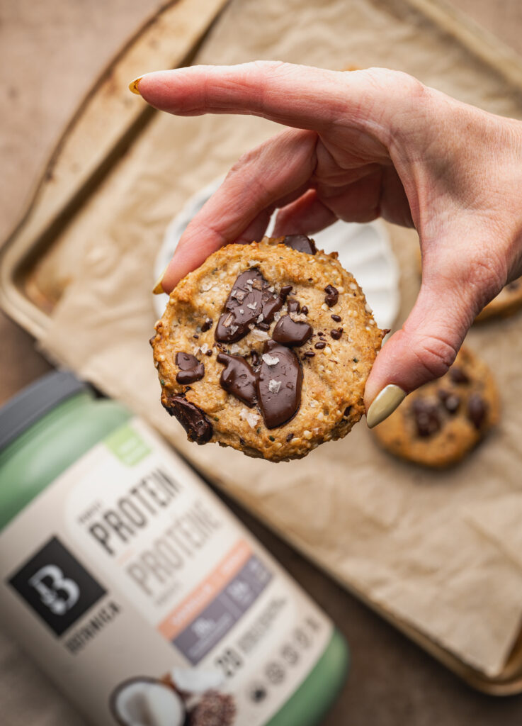 hand holding chocolate chip protein cookie in focus with container of plant based protein powder out of focus on the counter