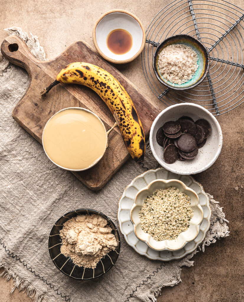 protein cookie ingredients on a cutting board such as runny tahini, banana, hemp seeds, chocolate chunks and plant based protein powder