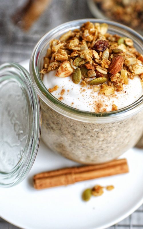 a glass jar filled with chia pudding. Granola is placed on top a cinnamon stick is placed on the plate below the jar.