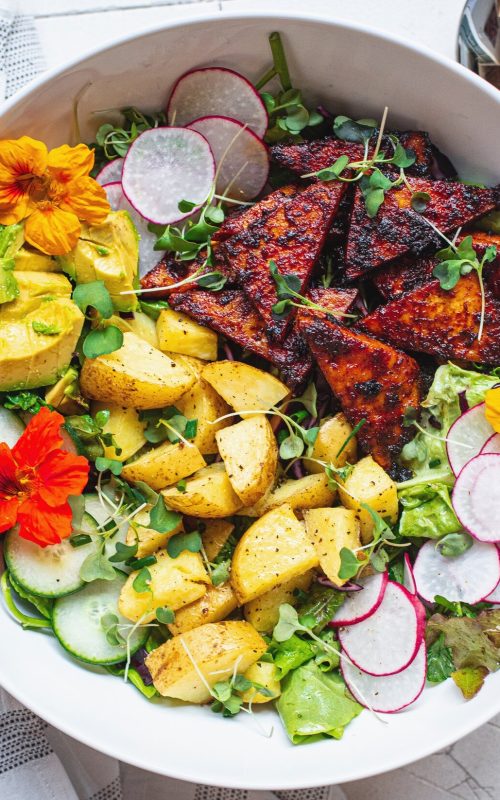 A beautiful salad bowl filled with colourful ingredients such as thinly sliced raddish, roasted potato, edible flowers, oven roasted bbq tofu, cubed avocado, sliced cucumber and micro greens