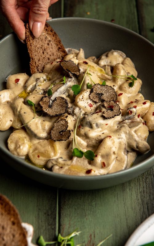 A close up image of Vegan BLACK TRUFFLE GNOCCHI pasta in a bowl. Thinly sliced Black truffle mushrooms are delicately placed on top.