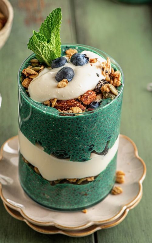 vegan yogurt and green chia pudding layered in a clear glass with granola blueberries on top. sitting on a scallop trimmed plate with gold rim and a fresh sprig of mint on top.