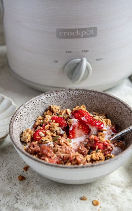 A bowl of strawberry rhubarb oatmeal with fresh slices of strawberries and a crockpot in the background.