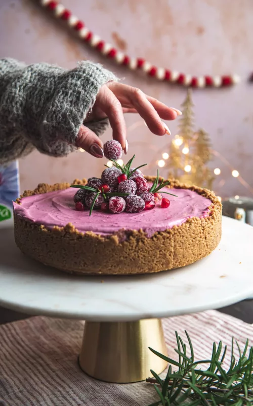 pink cheesecake with sugared cranberries and festive decorations. person placing sugared berry on top of cake