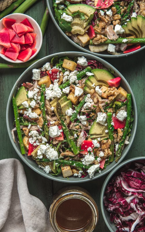 two bowls of a colourful salad filled with various vegetables, feta cheese and tofu. Loads of avocado and a hand adding some salad dressing to one of the bowls